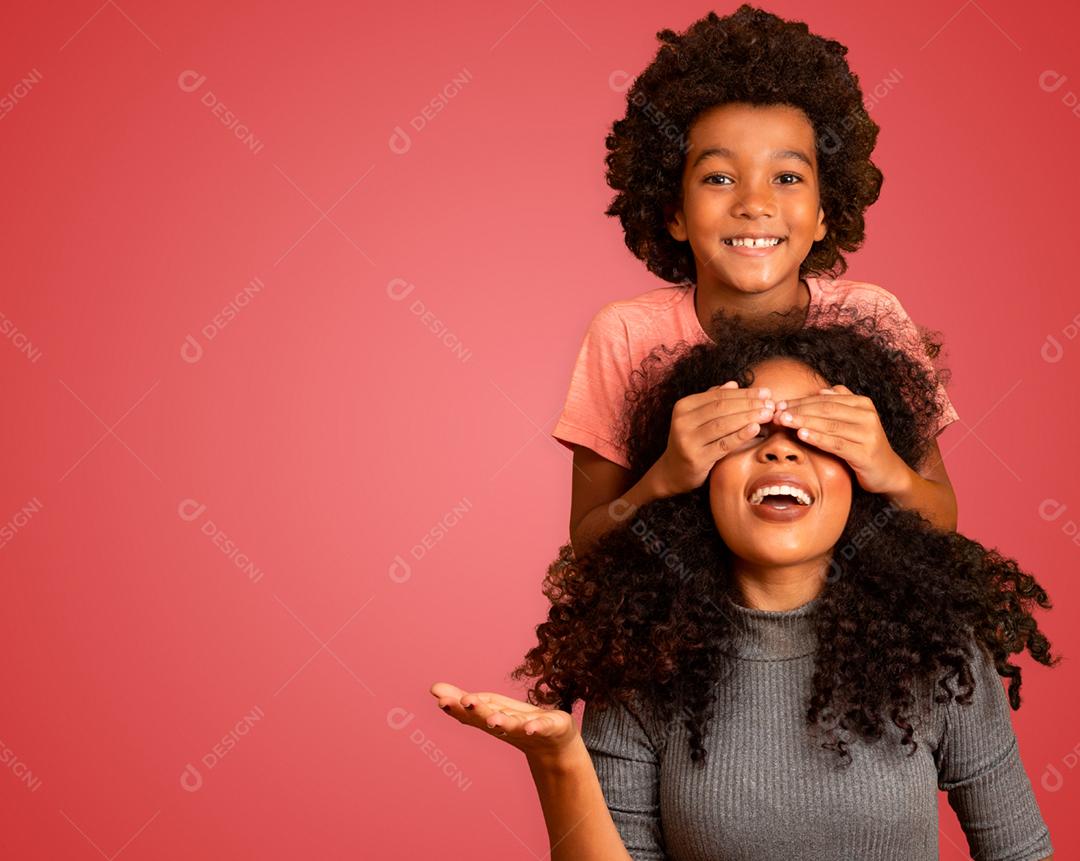 Menino afro-americano segurando os olhos fechados da mãe. Fundo vermelho. Dia das Mães. família brasileira.