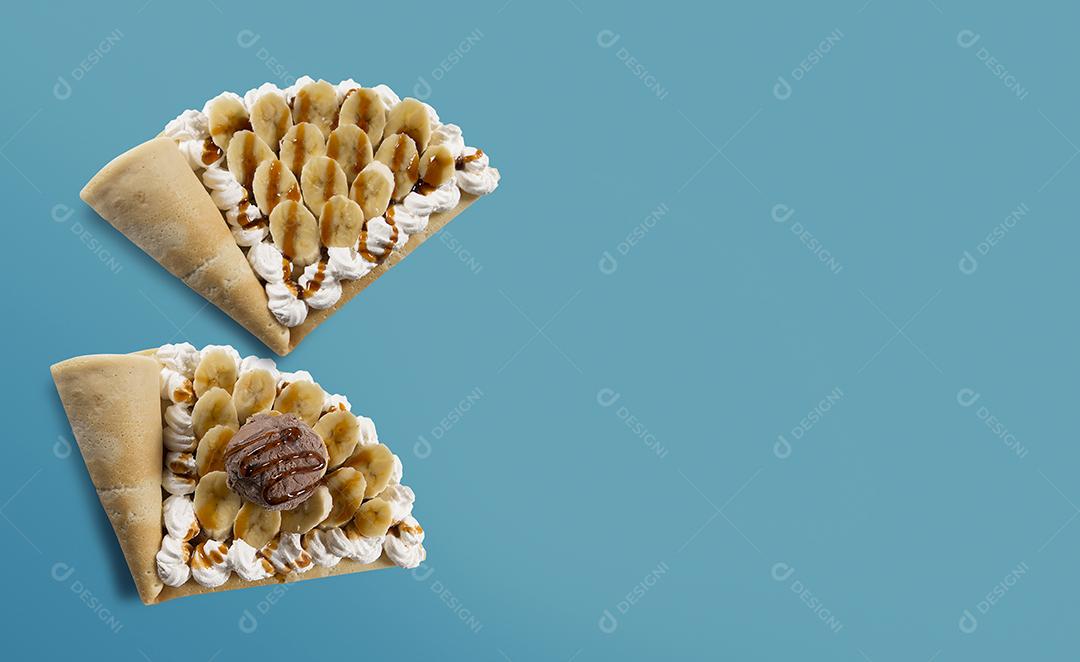 Vista superior de crepe ou panqueca fina sabor banana com sorvete de chocolate, pão de caramelo e mel com chantilly em fundo azul.