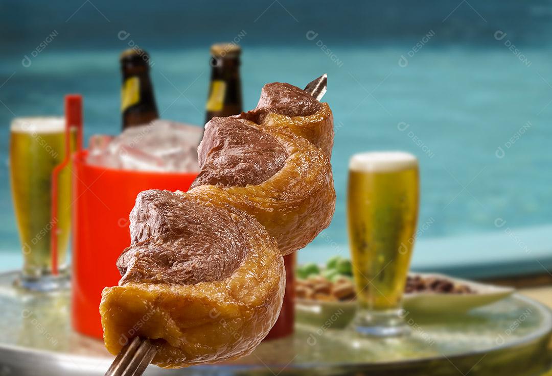 Picanha de churrasco brasileiro em espetos com cervejas turva e piscina ao fundo.