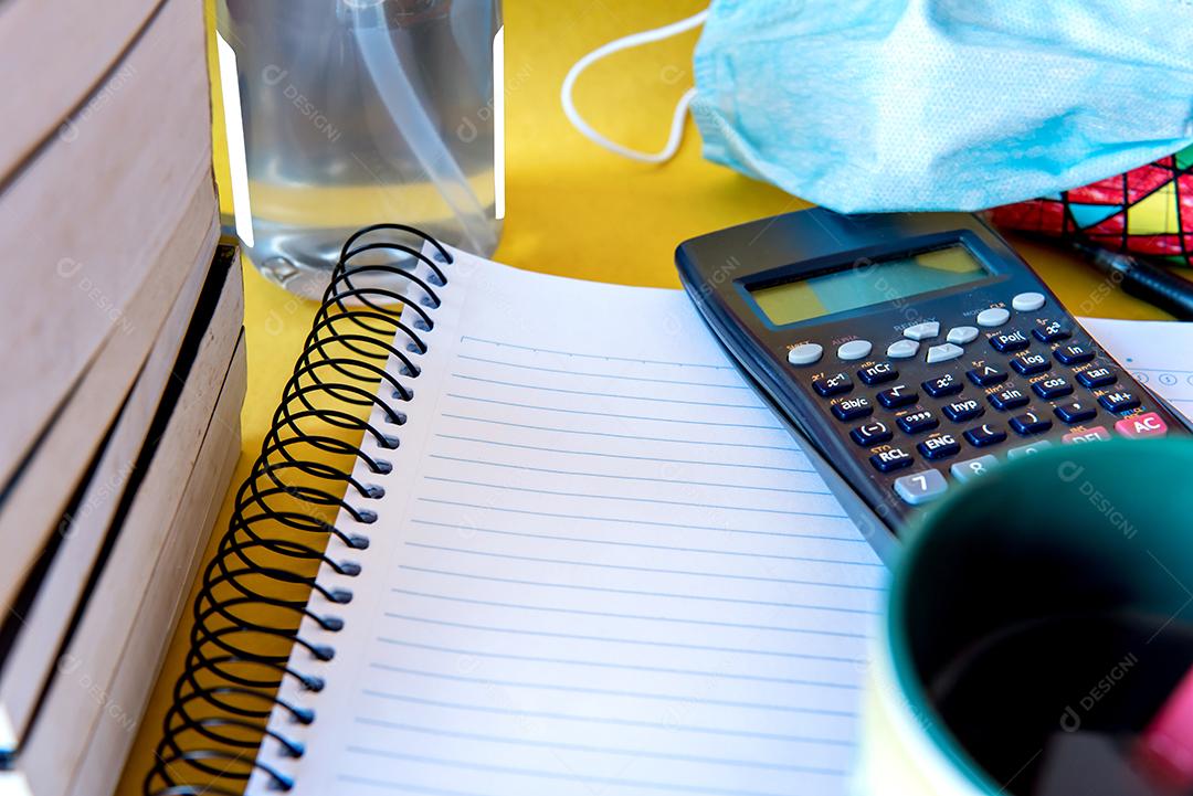 Mesa bagunçada com notebook, calculadora e livros. Educação, álcool gel e máscara, prevenção para COVID-19.